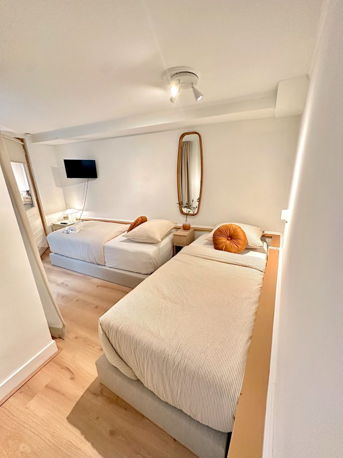 Cozy Twin Room A&A PAtio suite twin room. Lichte kamer met twee bedden, een televisie en een ronde spiegel aan de muur.